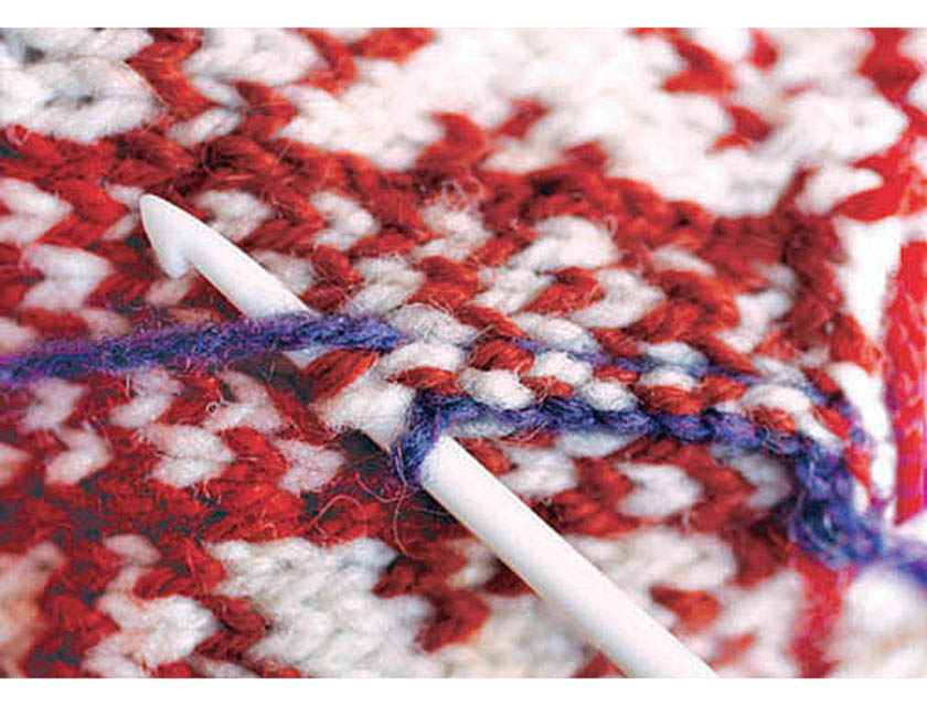 Close up showing the use of a crochet hook to secure steek stitches with blue colored wool on red and white pattern