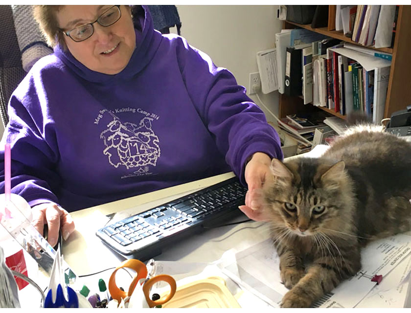 Tami Robus in purple Knitting Camp sweatshirt at her desk, petting a Maine Coon cat lounging in front of her keyboard