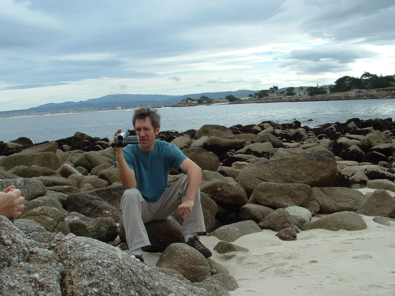 Cully Swansen sitting on gray rocks with Pacific ocean in background, filming with a video camera
