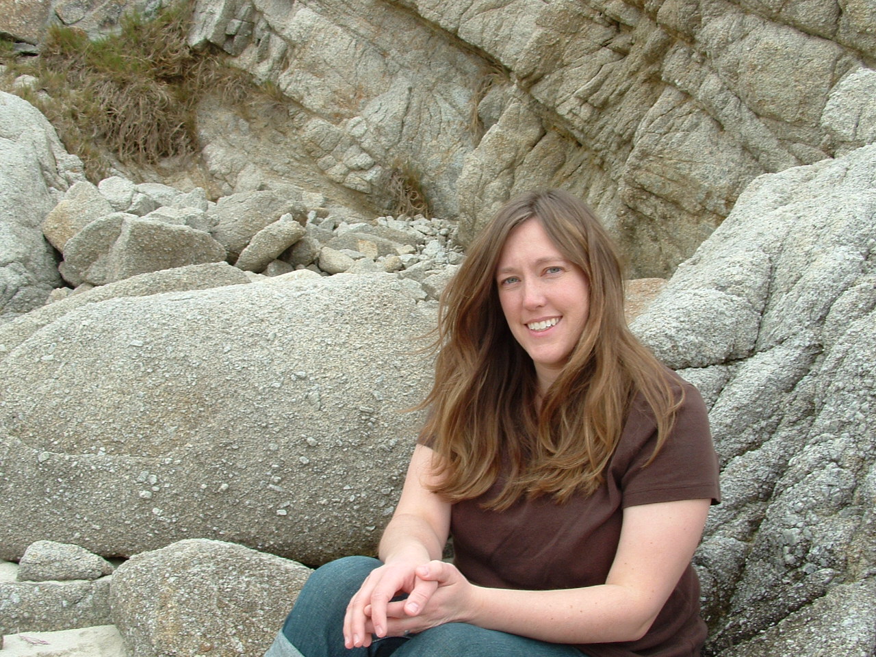 Michelle, a woman in brown shirt, sitting against gray rocks near the Pacific Ocean